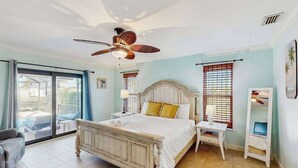 Une chambre à coucher comprenant un lit, un ventilateur de plafond, une table de chevet, une chaise et une fenêtre avec des stores.