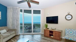 Una sala de estar con un sofá, dos mesas redondas azules, un televisor, un reloj y vista al océano a través de un ventanal corredero.