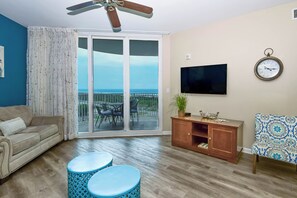 A living room with a sofa, two blue round tables, a television, a clock, and a view of the ocean through a sliding glass door. - 2BR~ Lagoon Pool~ Palms of Destin 21211 (Destin)