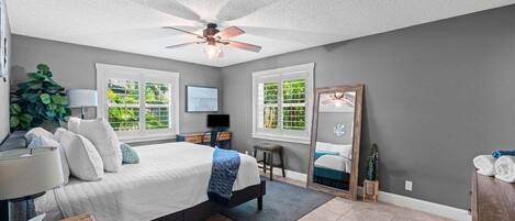 A bedroom with a bed, a desk, a chair, a ceiling fan, and two windows.