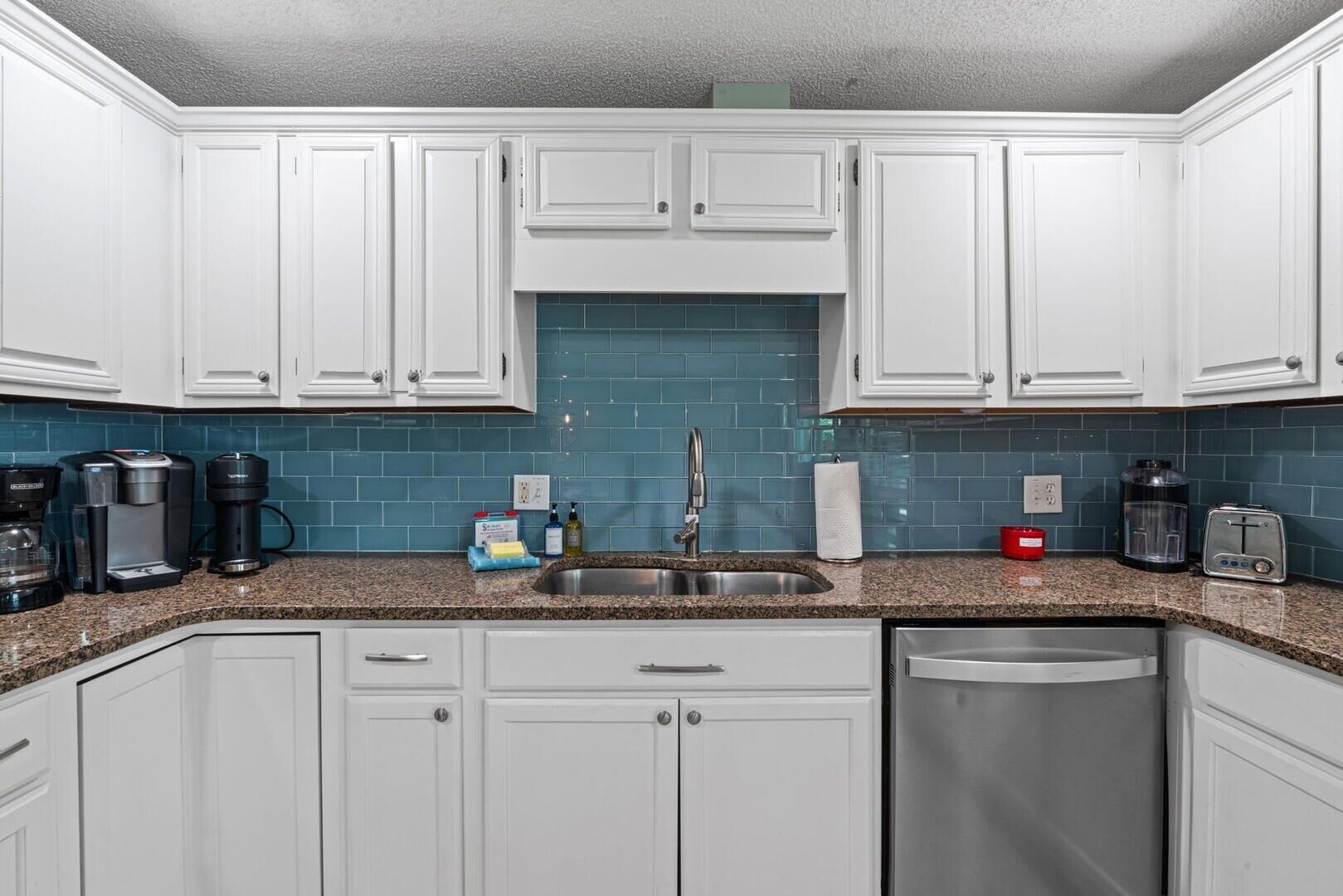 A kitchen with white cabinets, a blue backsplash, a stainless steel sink, and a dishwasher.