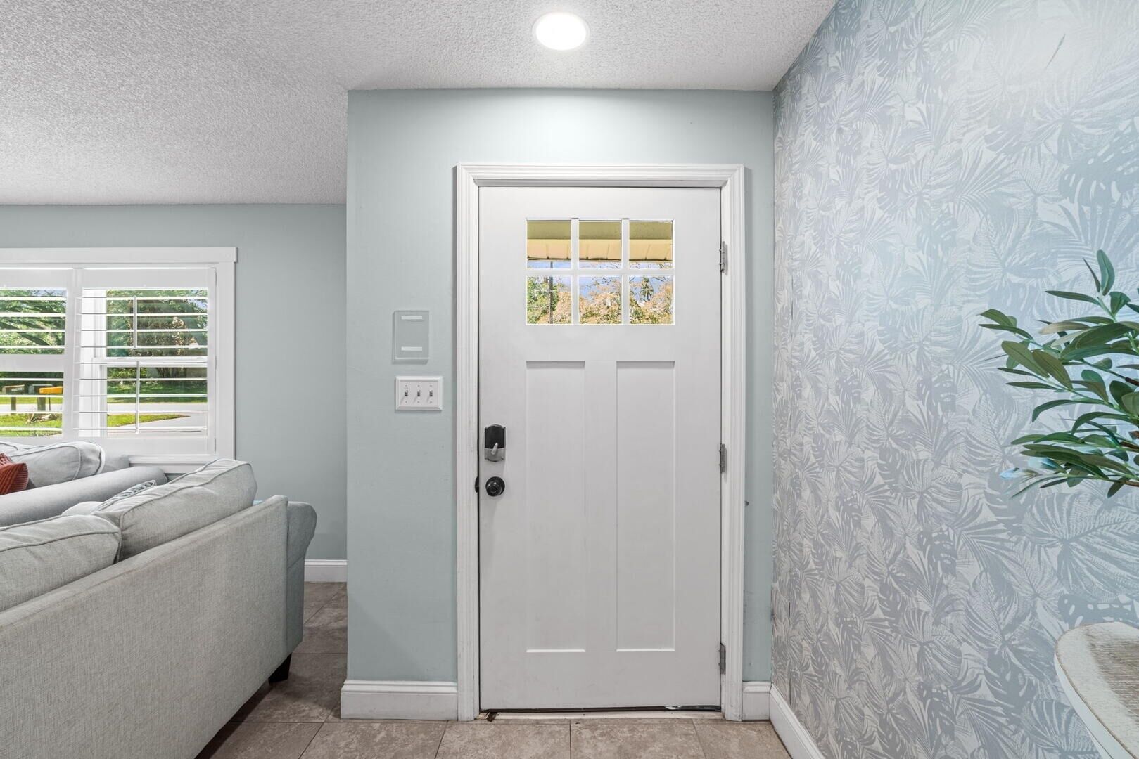 A hallway with a white door, a window with blinds, a grey sofa, and a patterned wall.