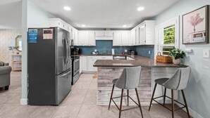 A modern kitchen with white cabinets, a stainless steel refrigerator, a granite countertop, and bar stools. - Vilano Beach I Ocean - Private Pool - Fire Pit 3 Bedroom Home (St. Augustine)