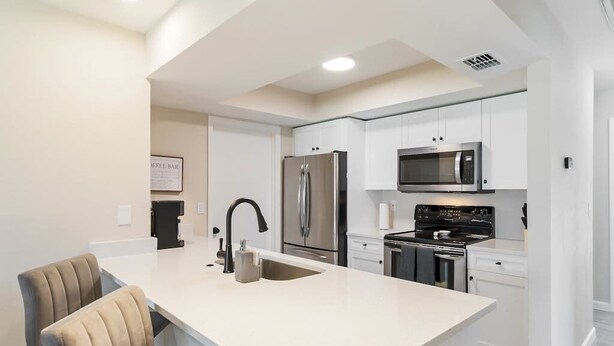 A modern kitchen with white cabinetry, stainless steel appliances, a central island with seating, and a sign indicating a coffee bar. - Centrally located duplex with on-site pool, firepit, patio, & BBQ - dog-friendly (Cape Coral)
