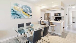 A modern kitchen with white cabinetry, stainless steel appliances, and a glass dining table with four chairs. - Centrally located duplex with on-site pool, firepit, patio, & BBQ - dog-friendly (Cape Coral)