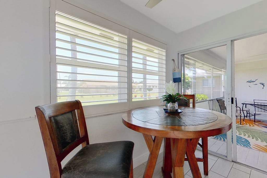 Una habitación con una mesa redonda de madera, una silla, una ventana con persianas y un portón corredizo de vidrio que da a un área exterior.