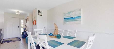 A dining area with a white table and chairs, a sailboat decoration, and a painting of a beach scene.