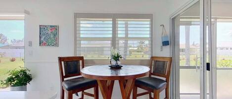 A dining area with a round table and four chairs, large windows with blinds, and a view of the outdoors.