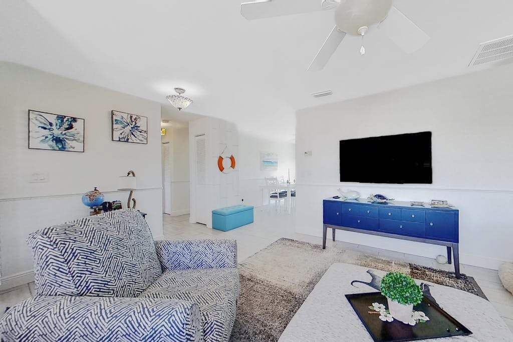 Una sala de estar moderna con un sofá, una mesa de centro con una planta, un otoman azul, un ventilador de techo blanco y un televisor de pantalla plana montado en la pared.