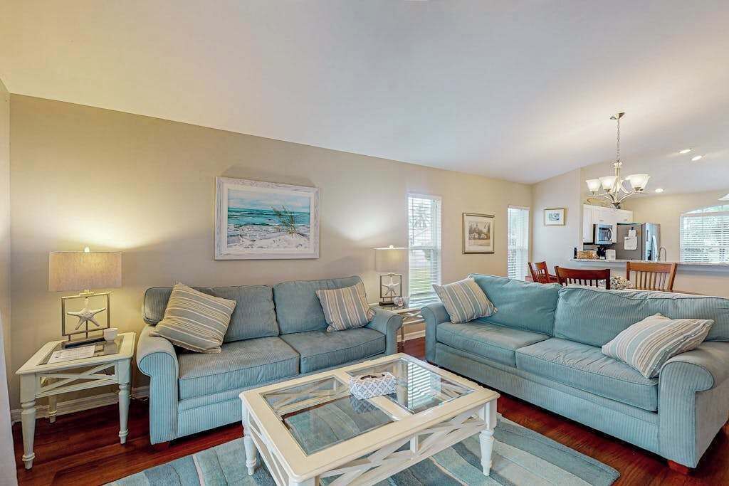 A living room with two light blue sofas, a glass coffee table, and a dining area with a chandelier.