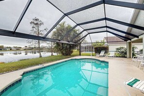 A swimming pool with a glass enclosure, surrounded by a grassy area and a body of water. - Waterside Home with Private Covered Pool & Patio - Golf Courses Nearby (Fort Myers)