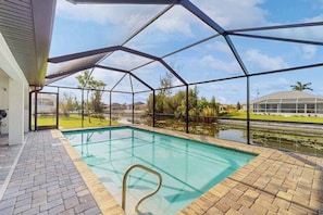 A swimming pool with a clear view of the surrounding landscape, a covered patio area, and a brick pathway. - Spacious Canal-Front home with private pool, lanai, Game Room (Cape Coral)