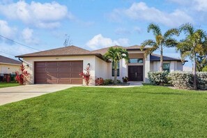 A single-story house with a garage, a front lawn, and a well-maintained garden with palm trees and shrubs. - Spacious Canal-Front home with private pool, lanai, Game Room (Cape Coral)