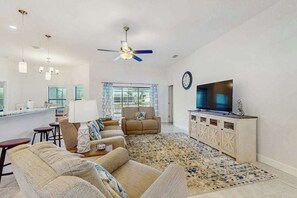 A living room with a sofa, a TV, a ceiling fan, and a kitchen area with a bar. - Spacious Canal-Front home with private pool, lanai, Game Room (Cape Coral)