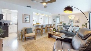 Una sala de estar moderna con un sofá, una mesa de centro y sillas, un comedor con mesa y sillas, y una cocina al fondo.