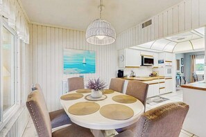 A dining area with a round table, chairs, a hanging light fixture, a painting, and a kitchen in the background.