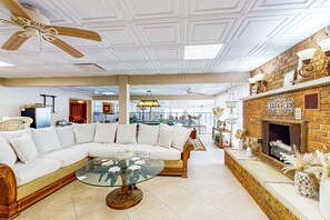 A modern living room with a sectional sofa, a glass coffee table, a fireplace, and a ceiling fan. - Luxurious single-level retreat w/ indoor pool, game room, and modern comforts (Fort Myers)