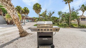 Ein tragbarer Grill mit mehreren Brennern auf einem Kiesgrund, umgeben von Palmen und Gebäuden.