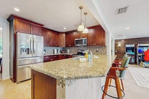 A modern kitchen with granite countertops, stainless steel appliances, wooden cabinets, and a bar area with stools. - Coastal home with private pool, arcade games, and firepit (Fort Myers)