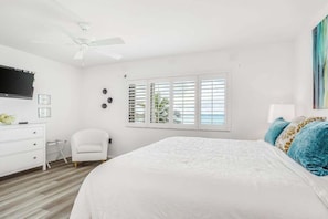 Ein Schlafzimmer mit einem Bett, einer weißen Kommode, einem Sessel, einem Deckenventilator und einem Fenster mit Rollläden.