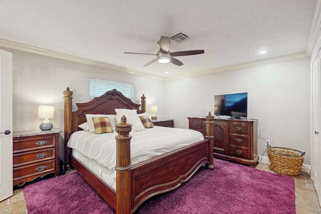 Een slaapkamer met een houten bed, een plafondventilator, een televisie, een dressoir en een paars vloerkleed.