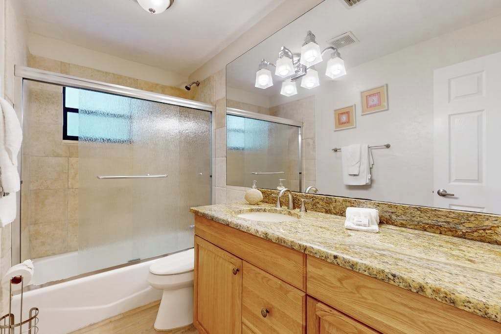 A bathroom with a glass-enclosed shower, a white toilet, a granite countertop sink, wooden cabinets, and a large mirror with lights.