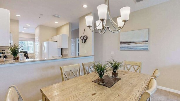 A dining area with a wooden table, chairs, a chandelier, and a kitchen in the background. - Upgraded Greenlinks Golf Villas Condo | All-New Appliances | Luxe Pool & Spa (Naples)