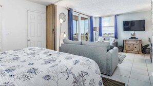 Une chambre à coucher avec un lit, un canapé gris, une télévision et une fenêtre avec des rideaux.