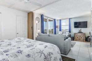 Ein Schlafzimmer mit einem Bett, einem grauen Sofa, einem Fernseher und einem Fenster mit Vorhängen.