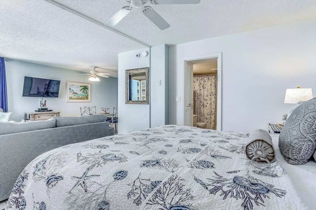 Ein Schlafzimmer mit einem Bett, einem Deckenventilator, einem Fernseher, einem Spiegel und einer Tür zu einem anderen Raum.