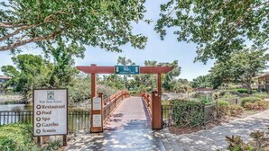 Un pont en bois mène à une zone fermée par une grille, et un panneau indique les équipements disponibles, tels qu’un restaurant, une piscine, un spa, un belvédère et un court de tennis.