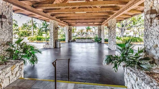 Une terrasse extérieure couverte, dotée de piliers en pierre, d’un sol en béton et de plantes en pot.