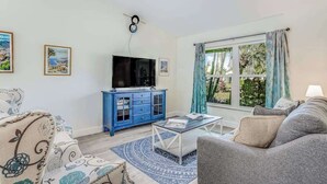 A living room with a sofa, a coffee table, a TV on a blue cabinet, and a window with curtains. - Beautiful home with private pool & lanai - near beaches & parks (Bonita Springs)