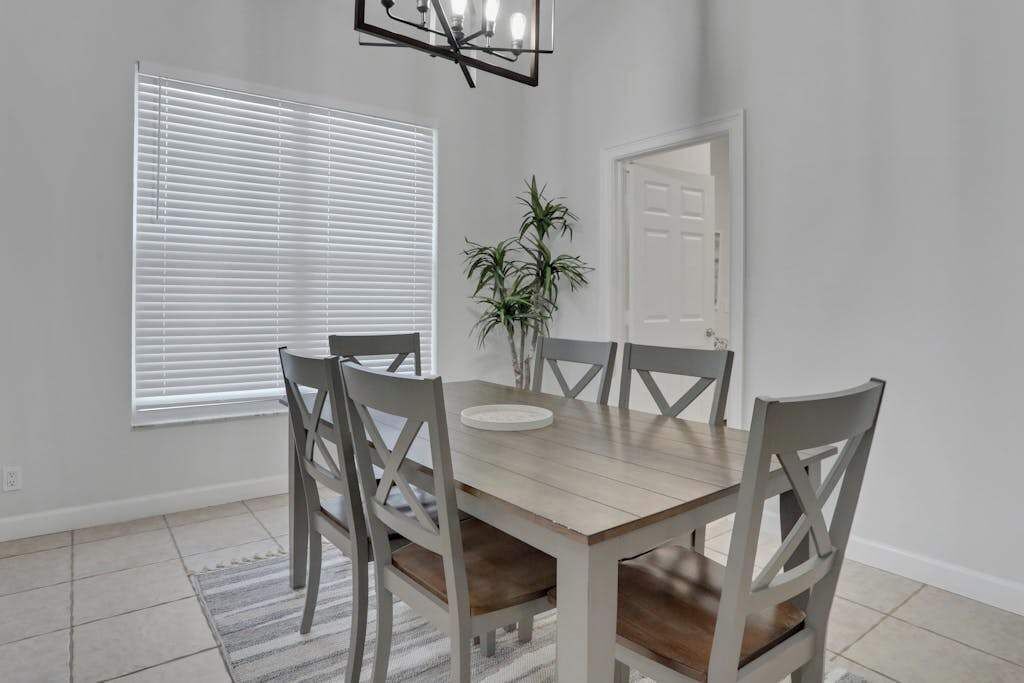A dining area with a wooden table, four chairs, a vase with plants, and a window with blinds.