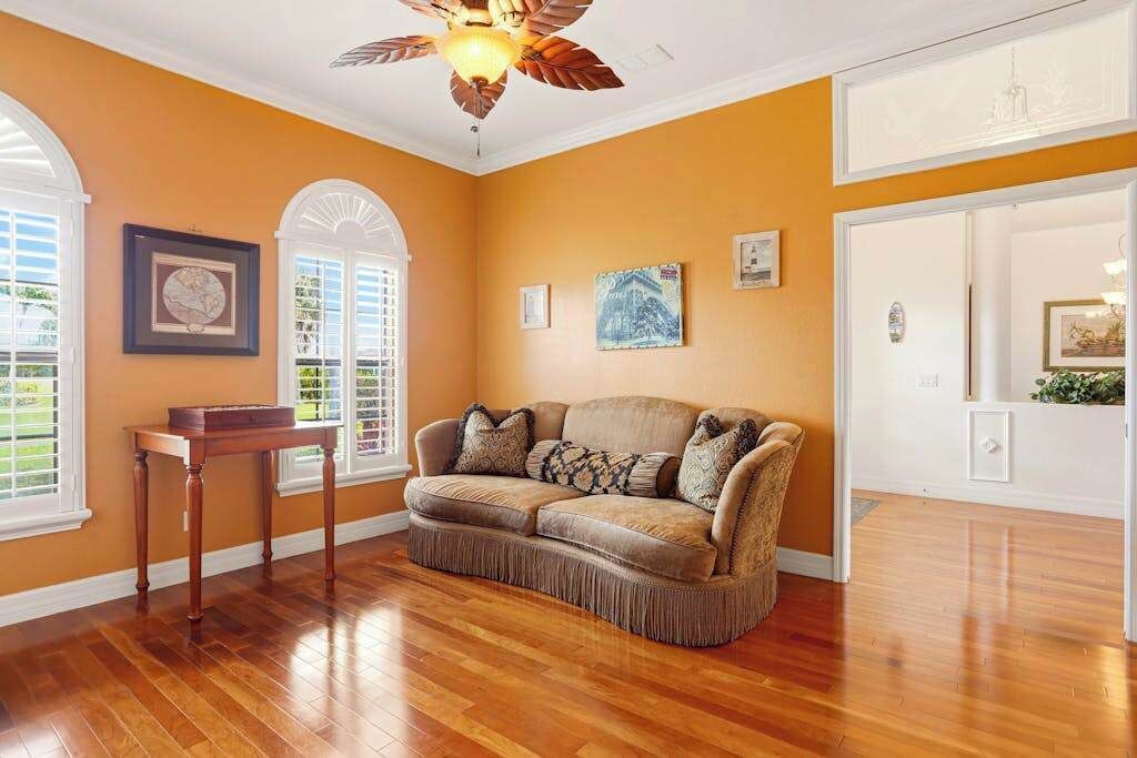 A room with a wooden floor, a brown sofa, a ceiling fan, and framed artwork on the walls.