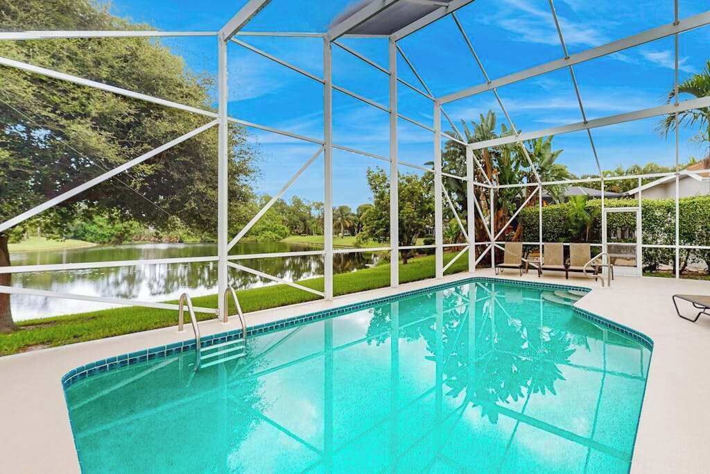 A swimming pool with a clear blue water, surrounded by a white fence, and a view of a lake and greenery.