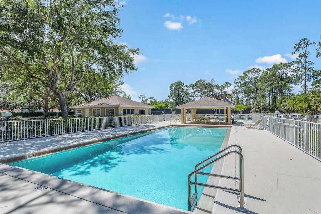 A rectangular swimming pool with a metal ladder, surrounded by a white fence, trees, and a gazebo in the background.