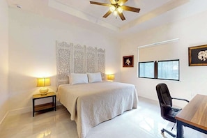 A bedroom with a bed, bedside lamps, a ceiling fan, a desk with a chair, and a window.