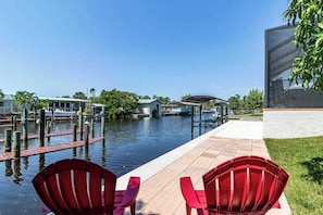 A waterfront view with a dock, a covered area, and red chairs on a brick pathway. - Spacious, dog friendly home with pool, on waterway within minutes to the Gulf (Bokeelia)