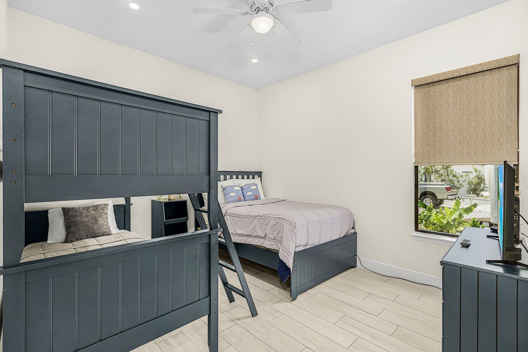 A bedroom with a bunk bed, a single bed, a desk, and a window with a view of greenery.