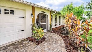 A residential house with a garage, a front door with a decorative glass panel, and a landscaped garden with various plants. - Canal-front home w/ private pool, hot tub, dock & kayaks/paddleboards -1 dog OK (Cape Coral)