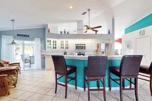 A kitchen with a central island, bar stools, a ceiling fan, and a sign reading 'RATHY'S CAFE'. - Canal-side gem with screened pool, boat dock & Gulf access - near dining (Cape Coral)
