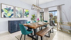 A dining area with a wooden table, teal chairs, and a bench. There are framed botanical prints on the wall, a hanging light fixture, and a view through the window. - Dog-friendly (hypoallergenic), Heated pool, Game Room, Smart T.V.'s and More (Cape Coral)