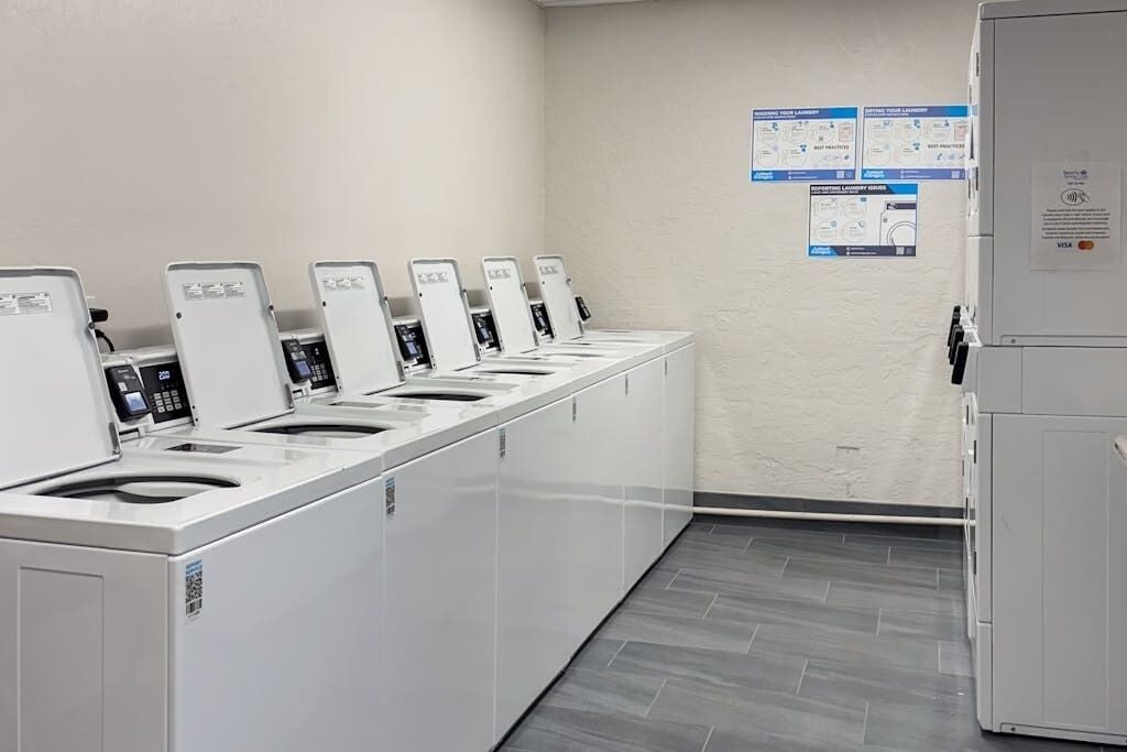 A row of white self-service laundry machines with control panels, a tiled floor, and informational posters on the wall.