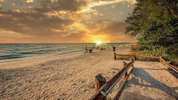 Une plage avec une promenade en bois menant à l’eau, un coucher de soleil en arrière-plan et des gens sur le sable.