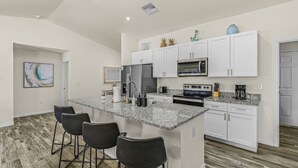 Une cuisine moderne avec des armoires blanches, un comptoir en granit, un réfrigérateur en acier inoxydable, un four à micro-ondes et une cuisinière. On y trouve trois tabourets de bar et un tableau accroché au mur.
