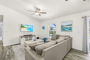 A modern living room with a sectional sofa, a flat-screen TV, a ceiling fan, and artwork on the walls. - Cape Coral Retreat | Heated Pool, 2 King Suites & Dog-Friendly Game Room (Cape Coral)