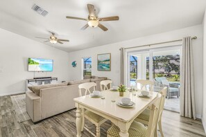 A modern living room with a sofa, dining table set, ceiling fans, and artwork on the walls. - Cape Coral Retreat | Heated Pool, 2 King Suites & Dog-Friendly Game Room (Cape Coral)