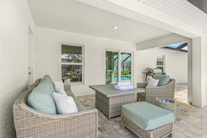 A covered outdoor seating area with wicker furniture and a table, a pool visible through the glass doors, and a ceiling with a textured finish. - Cape Coral Retreat | Heated Pool, 2 King Suites & Dog-Friendly Game Room (Cape Coral)