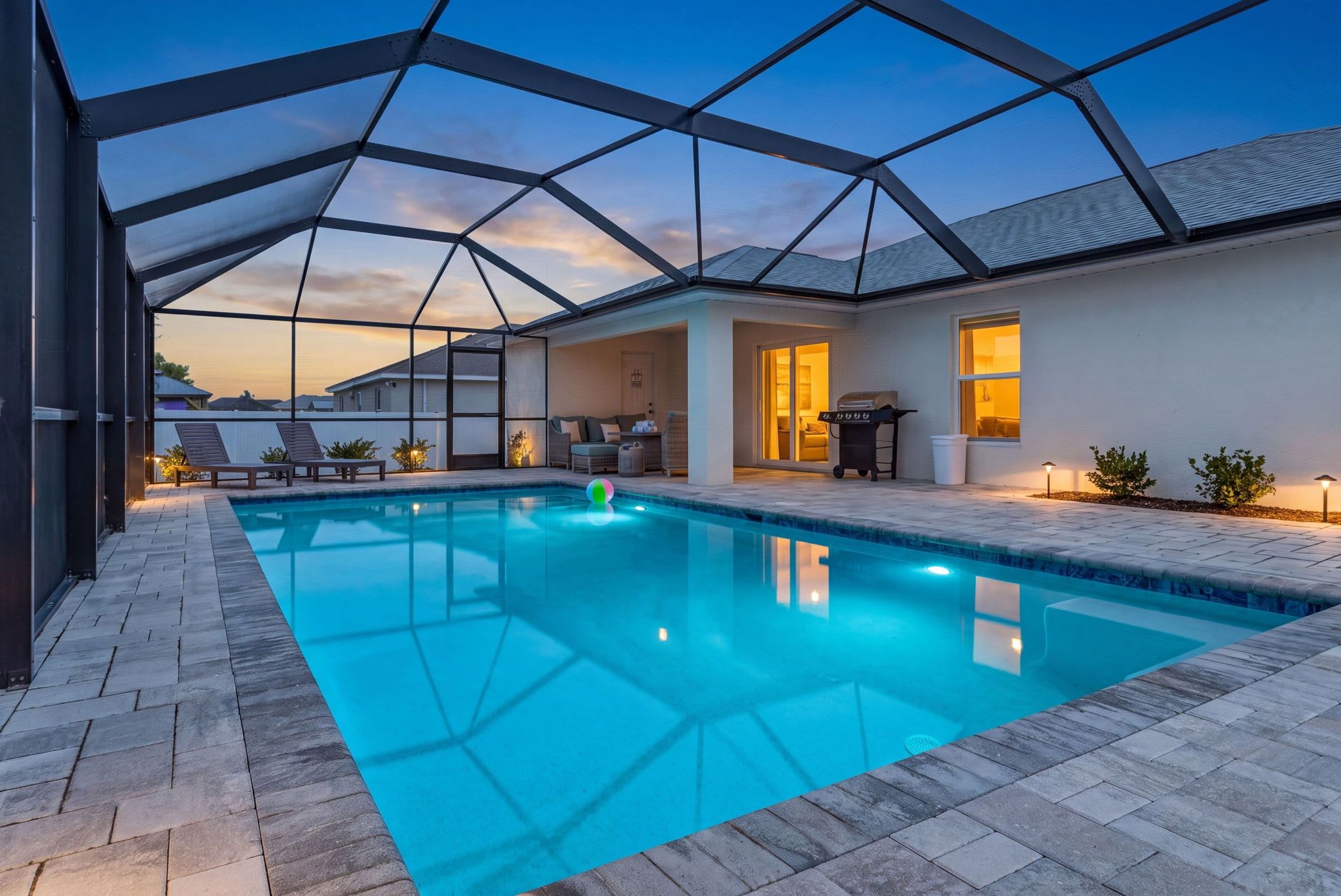 Una piscina con cerramiento de vidrio, un área de patio con asientos y vistas al atardecer.
